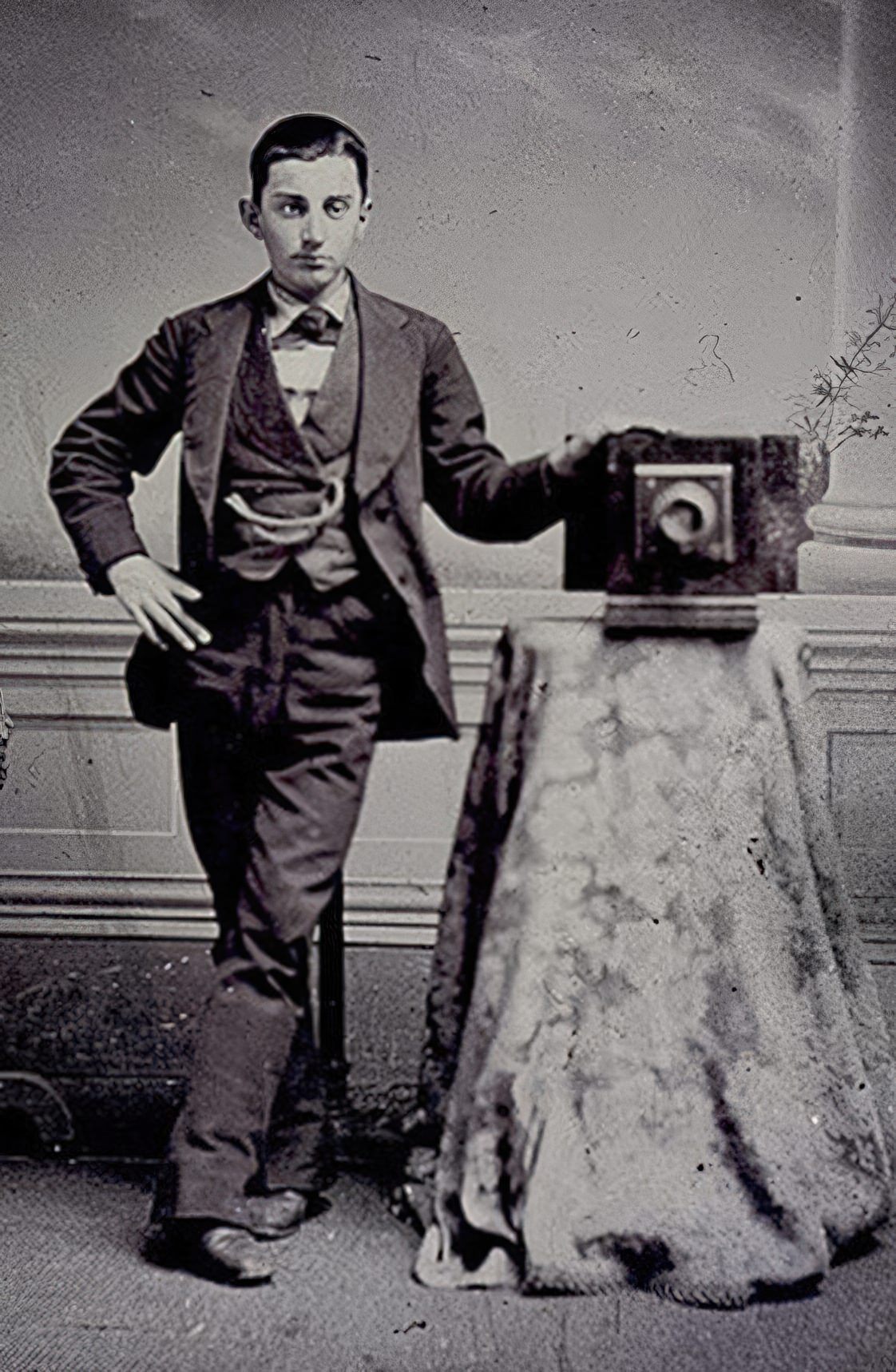 In this tintype a young man stands beside a table holding a camera.
