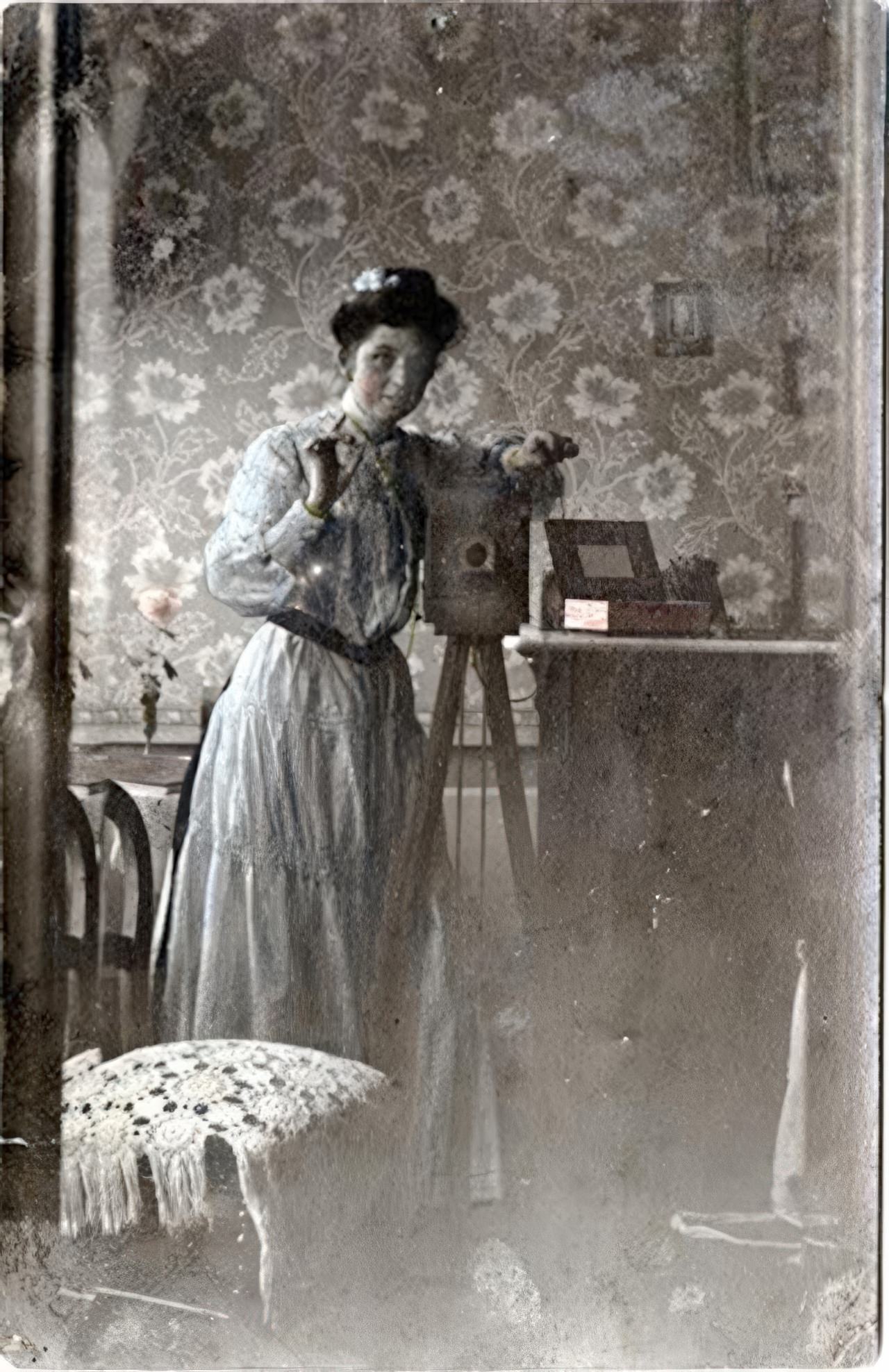 A young woman in a tinted blue dress and her camera are reflected in a large mirror. The left side shows a double image of a chair and the wall paper in the beveled edge of the mirror. This real photo postcard is marked "Rébublique Française" and "Carte Postale".