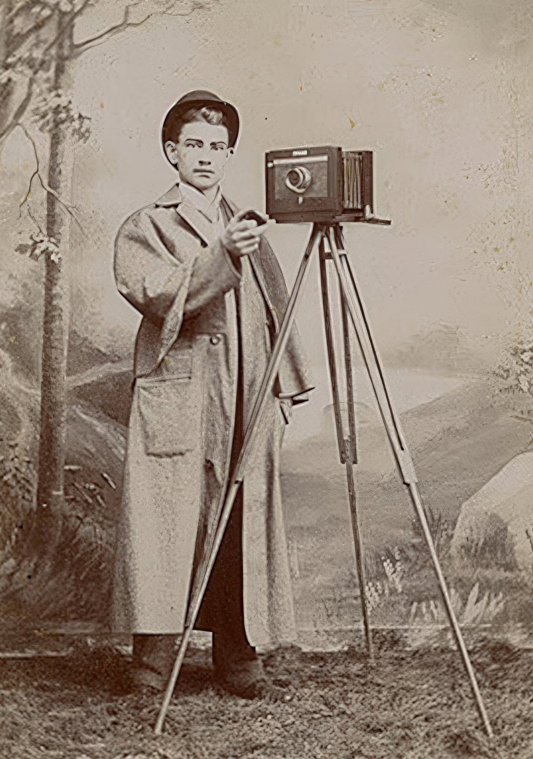 In this cabinet card a young man uncaps the lens of his tripod mounted camera to make an exposure. In spite of the caped coat, grass, and landscape background it was taken in a studio. The background is unusual because it seems to be painted directly on the wall rather than on canvas. The landscape has been painted over the baseboard of the wall. The photograph is by Whitley of Elmira, NY.
