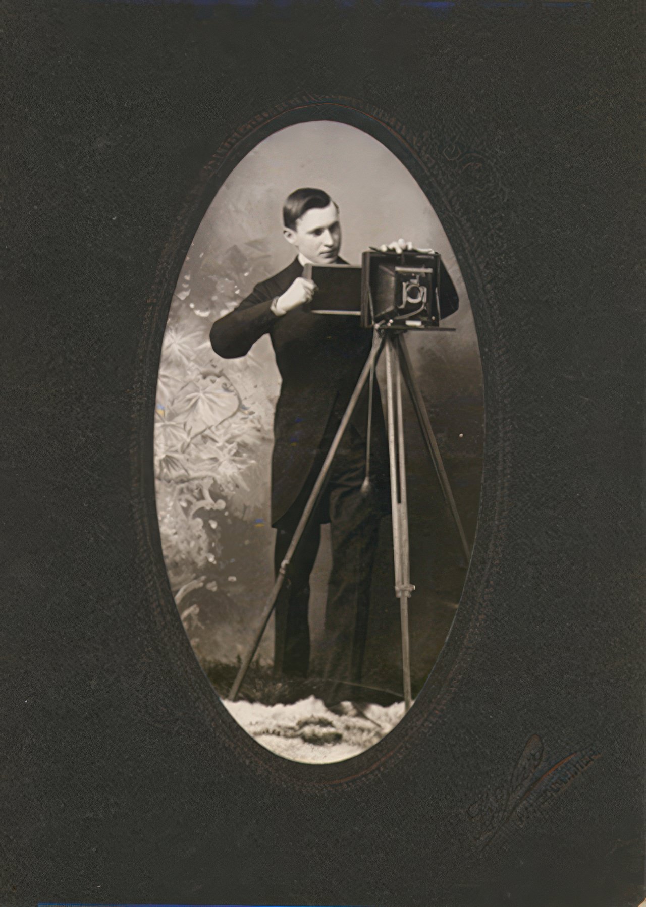 A well dressed young man inserts a film holder into a folding plate camera on a tripod in this oval cut photograph on a heavy black mat. The photographer's name is embossed in hard to read script but the location, Hancock, Michigan is clear.