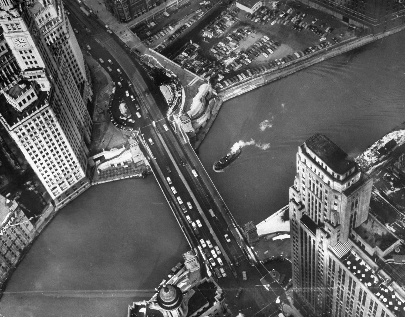Chicago's famous Wrigley Building looks like candy castle from a helicopter above spire. Building is split in two parts and a railroad track runs between them. Behind them is Chicago River, with Michigan Avenue bridge.