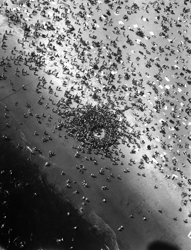 Beach accident, the near drowning of a Coney Island bather named Mary Eschner, draws knot of people. Reviving victim lies in center, attended by lifeguards. Some bathers (foreground) wave a helicopter as they run from water.