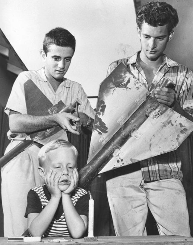 Four-year-old Mark Malone of Burbank sticks to old-fashioned firecracker as his buddies, Myron Lieberman, 18, center, of Burbank, and Leston Newbill, 17, of Burbank, prepare to launch three-stage rocket they built for July Fourth celebration. 1959.