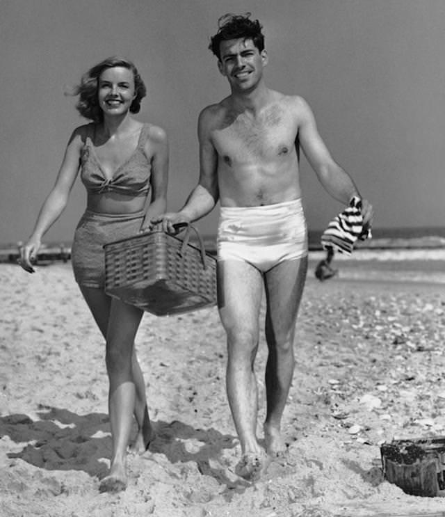 Circa 1950s: Couple walking with picnic basket on beach.