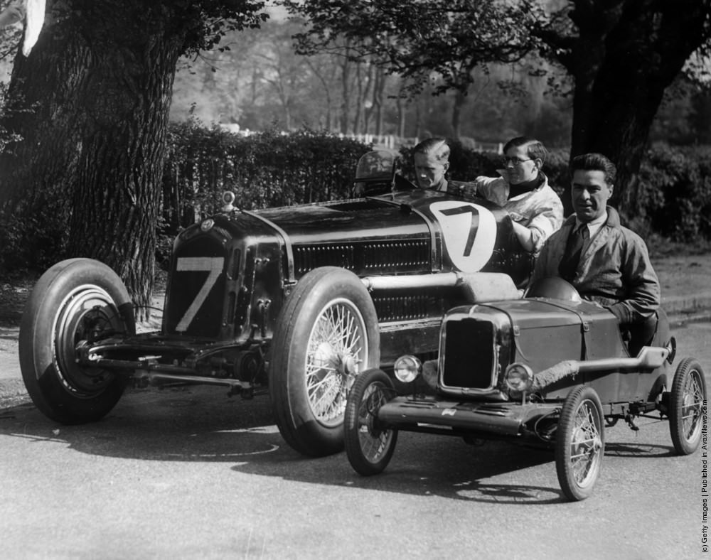 Dwarfed by a full-size car, Mr C W Cooper of Surbiton drives the miniature racing car which he built for his son John. The tiny vehicle is fitted with a 1.25 horsepower two-stroke engine, and can travel up to 52 miles per hour. 27th May 1935.