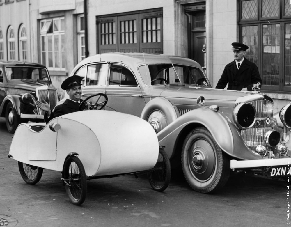 A tiny Velocar makes its way down the road in Hyde Park, London, dwarfed by the parked cars alongside. This pedal-powered “one manpower” vehicle is one small answer to the problem of petrol rationing. 5th November 1939.