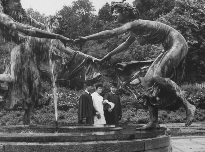 A trio of newly graduated students strolls soberly past a trio of figures dancing ring-around-a-rosy at a fountain