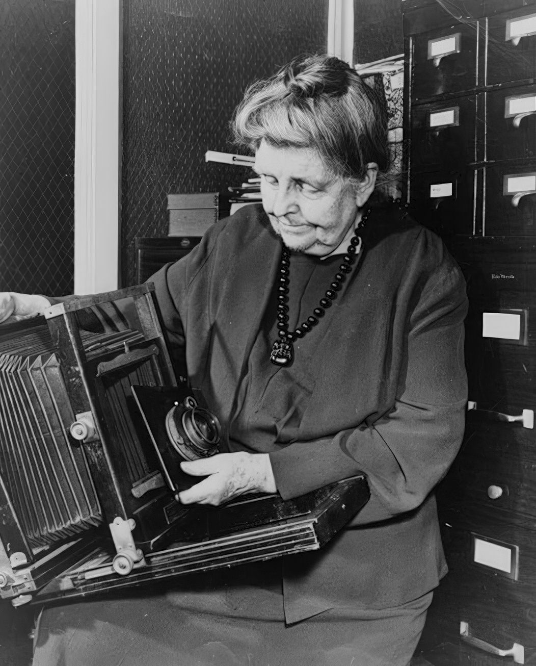 Frances Benjamin Johnston, three-quarter length portrait, holding and looking down at camera, facing slightly left, 1950.