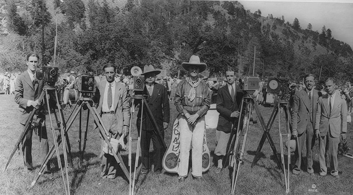 President Coolidge in cowboy outfit, standing in field with photographers; mountain in background, 1927.
