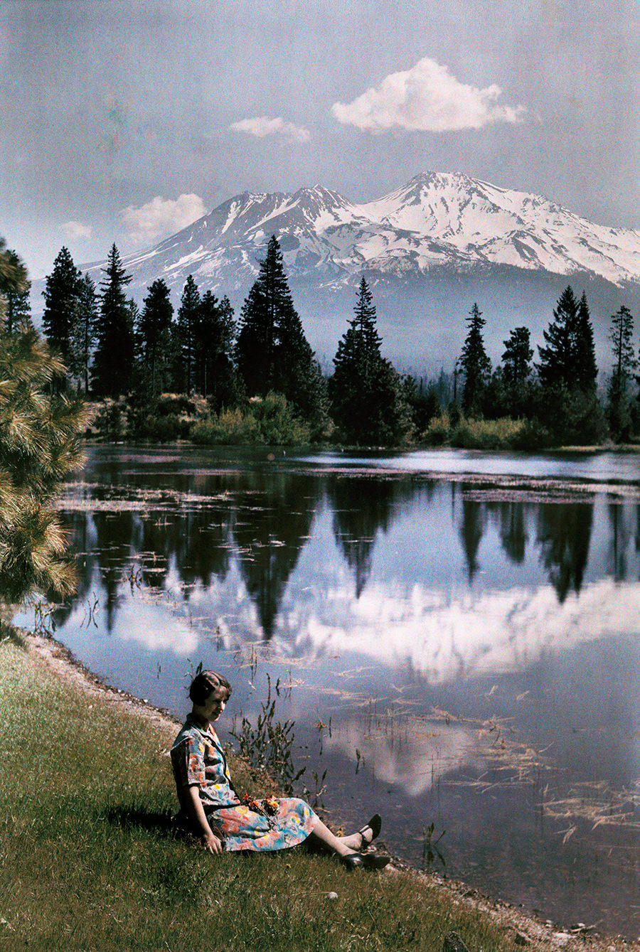 A girl sits by a lake with snow-capped mountains in the background, California, 1929.