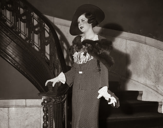 Model Jean Early poses on the staircase at a 1934 summer fashion show while completely decked out in an elegant suit with a fur cape and hat.