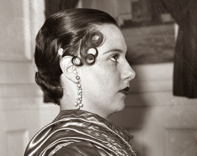 Looking very severe, a model shows off a distinctly 1930s hairstyle with curls above the eye and her hair pulled tightly back.
