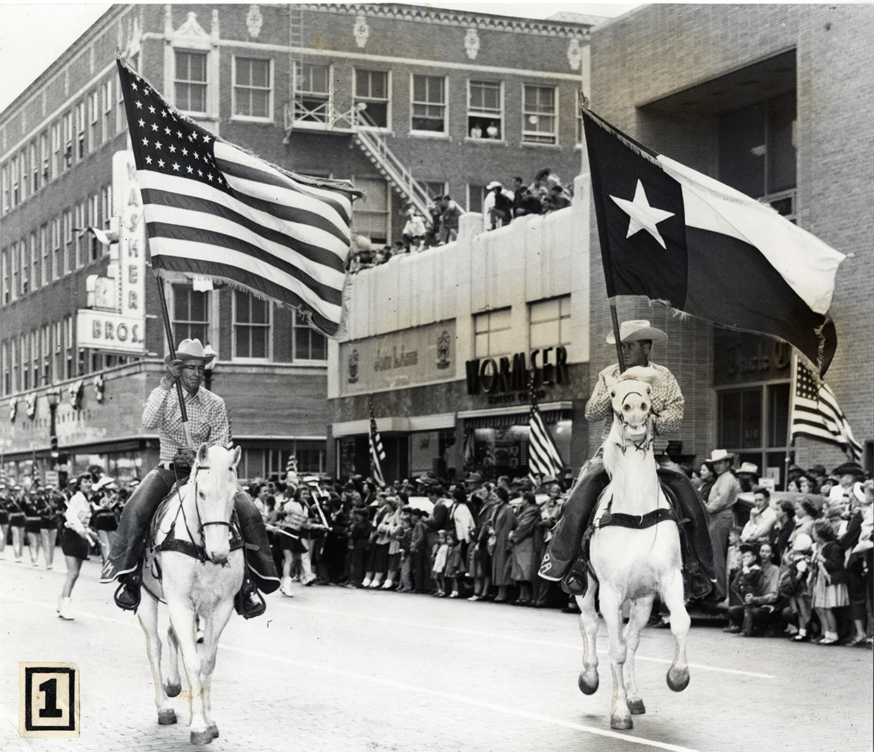 Two men on white horses in Stock Show parade carrying United States flag and Texas flag, downtown Fort Worth, Texas, 1953.