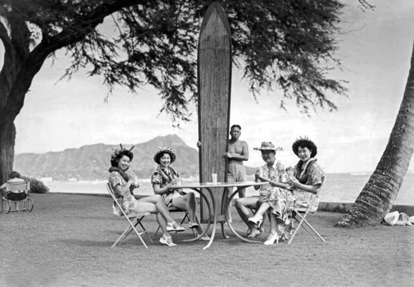 Four Japanese tourists pose at the Halekulani Hotel on Waikiki Beach with a native Hawaiian and his long surfboard, 1930.