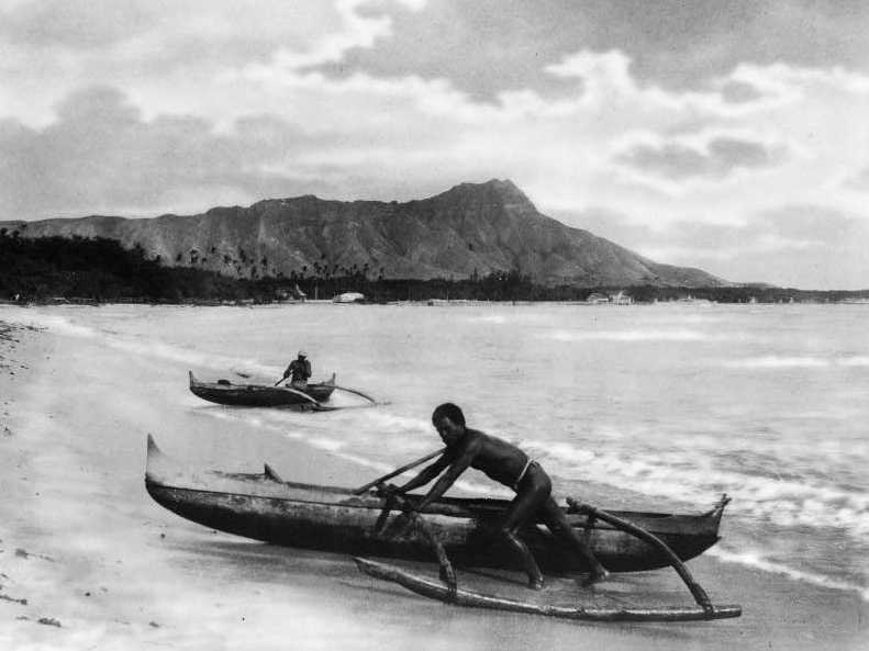 Two native Hawaiians with outrigger canoes at the shoreline in Honolulu, 1922.