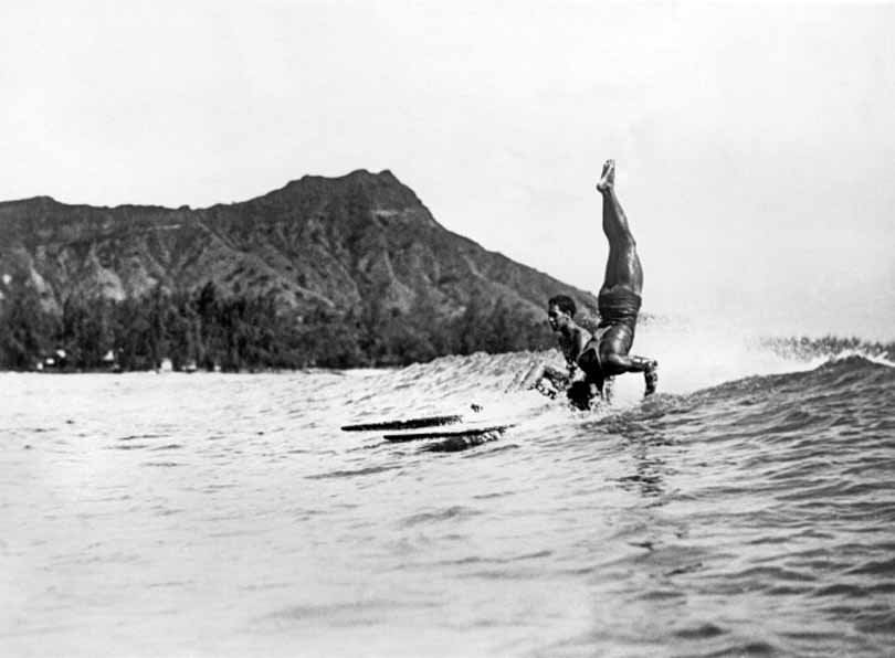 A surfer at Waikiki Beach stands on his head as he rides a wave into the shore, 1925.