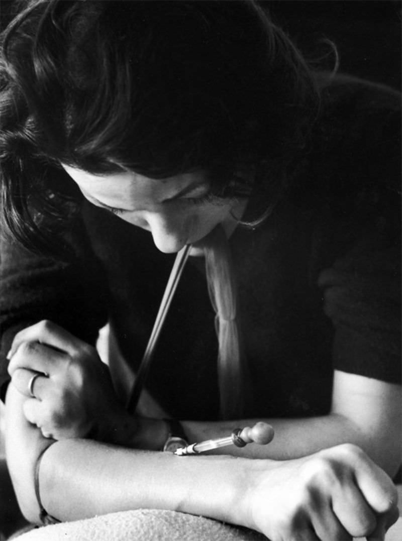 Karen with nylon stocking around her arm to make vein pop out as she uses hypodermic needle on an eyedropper which addicts call a "spike" to shoot up with heroin.