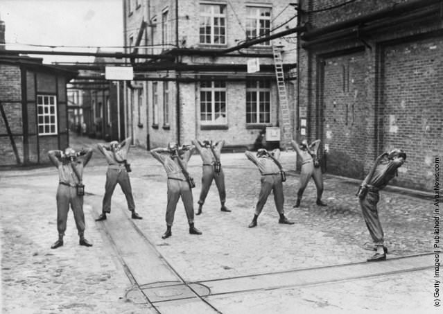 Men wearing gas masks exercising in a factory yard, 1935.