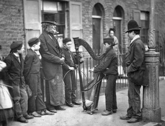 An Italian harpist entertaining children on the street, 1870s.