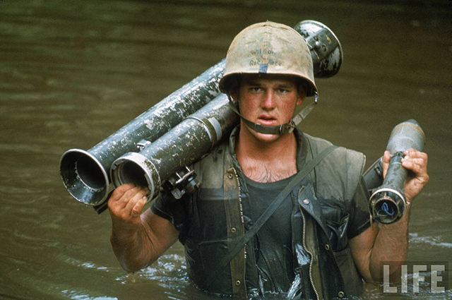 US Marine Phillip Wilson carrying rocket launcher across stream during fighting near the DMZ during the Vietnam War.