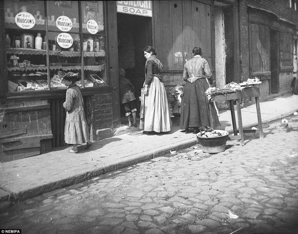 A young girl examines the window display of a city shop selling fresh veg, sunlight soap and sweets.