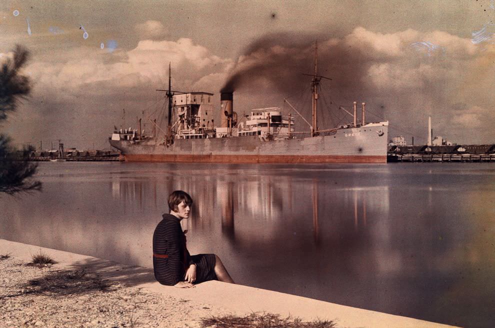 A woman sits on an edge by a harbor where a freighter boat approaches, 1930