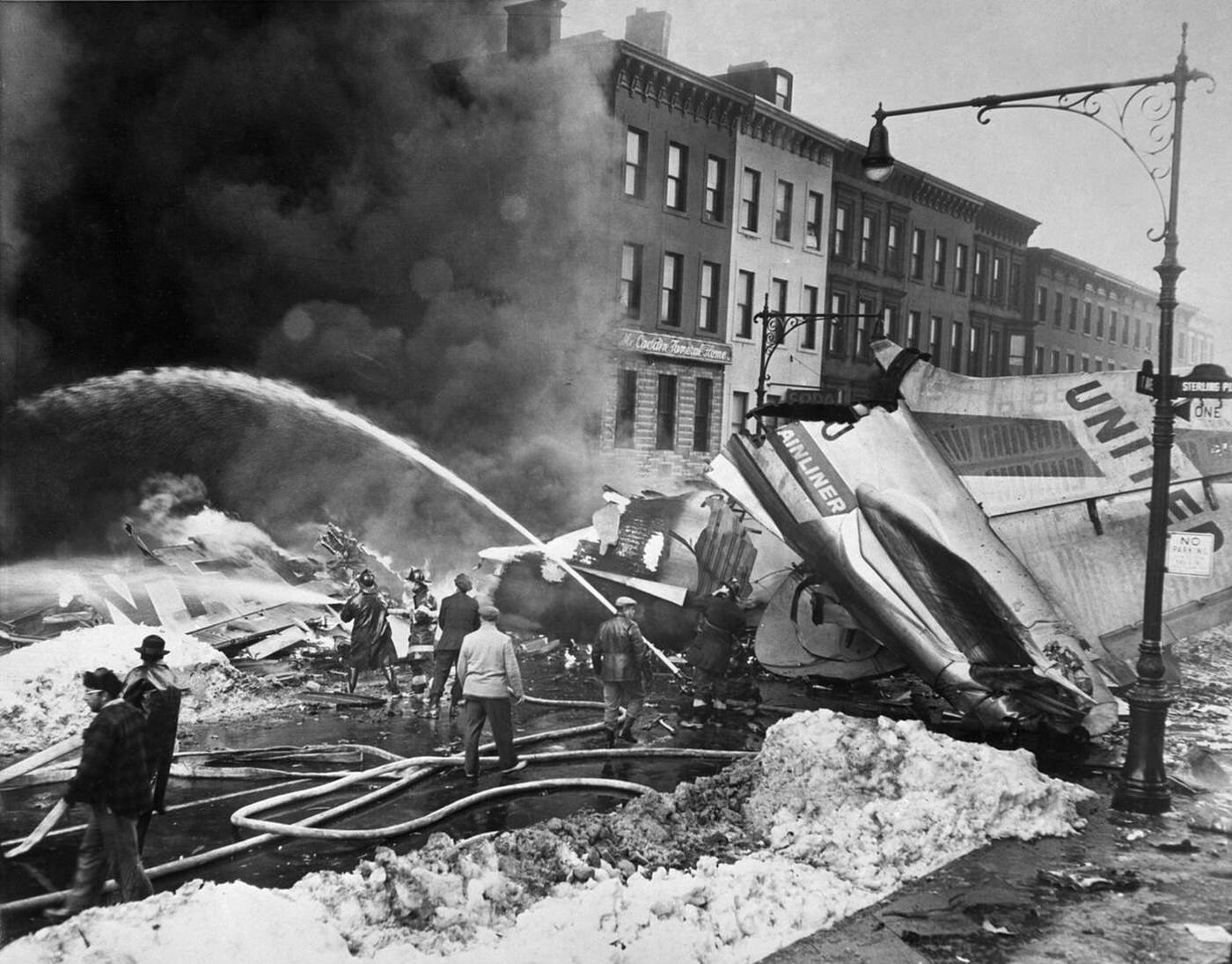 Firemen and rescue workers tackle the blaze in front of the tail-plane and wing of the United Airlines Douglas airliner, which lay in the middle of a street in residential Brooklyn after colliding mid-air with a TWA Constellation airliner; at least 132 people died in the disaster, December 1960.