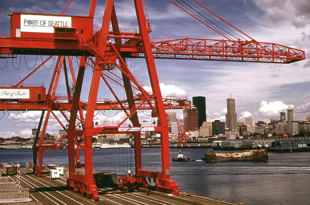 Port of Seattle freight handling equipment and city skyline, with an industrial waste barge under tow to landfill on the Snohomish River estuary.