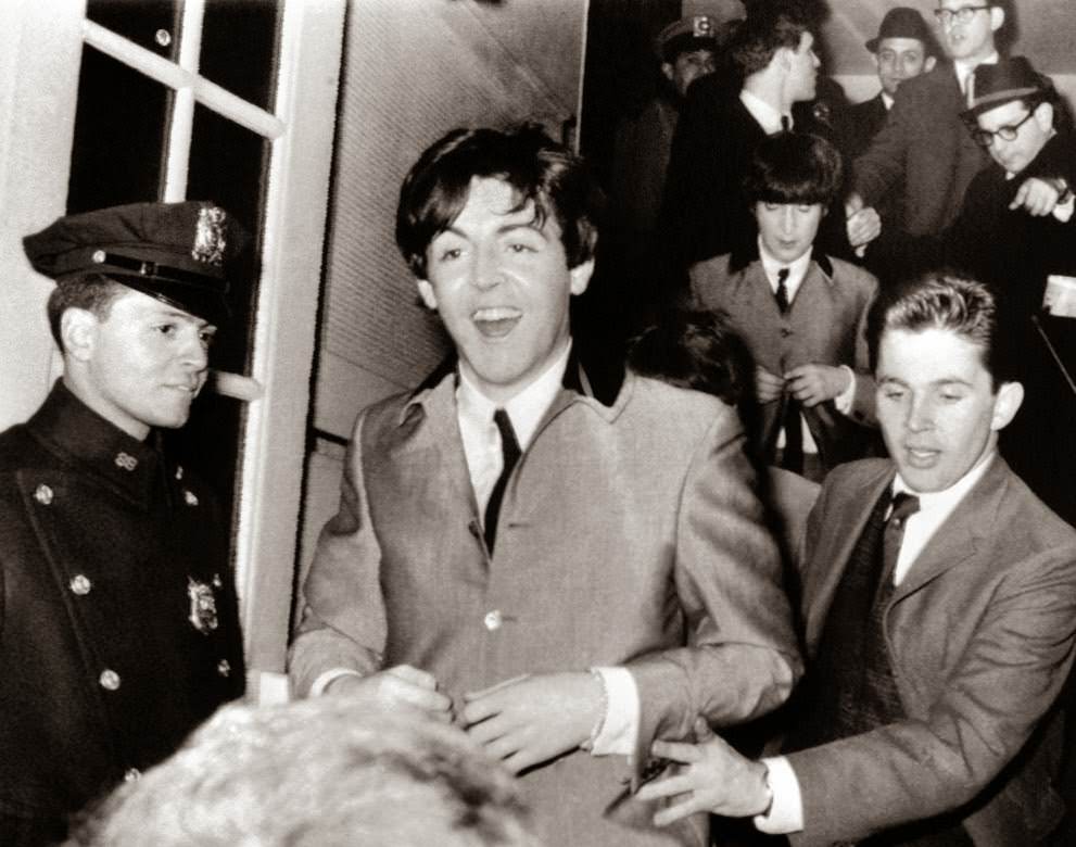 Beatle Paul McCartney flashes a smile as he rushes from New York's Carnegie Hall after two wild performances; band mate John Lennon is behind him, 1964.