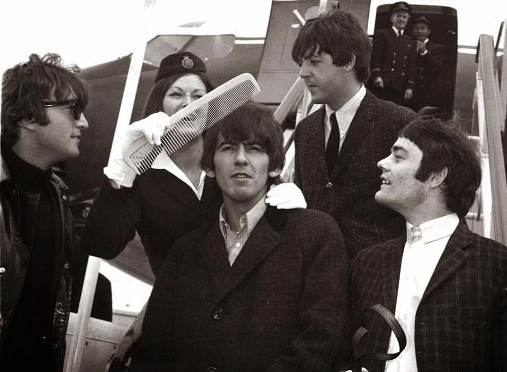 George Harrison gets the big comb treatment from BOAC stewardess Anne Creech, after their arrival at Windy airport in London; John Lennon, Paul McCartney and drummer Jimmy Nicol, who stood in for Ringo Starr for the trip to The Netherlands are also pictured, 1964.