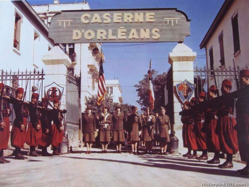 The Color Guard of the Women's Army Corps in North Africa is seen coming through the gate of the Caserne D'Orleans, a famous Mediterranean fortress, while the guard of honor of the First Zouave Regiment stands at attention.