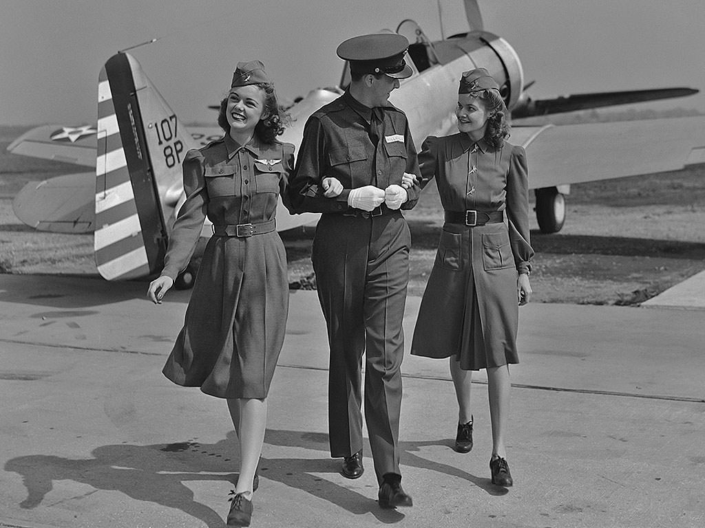A male flight attendant walks with his arms linked with two female flight attendants in front of a small plane in the 1940s.
