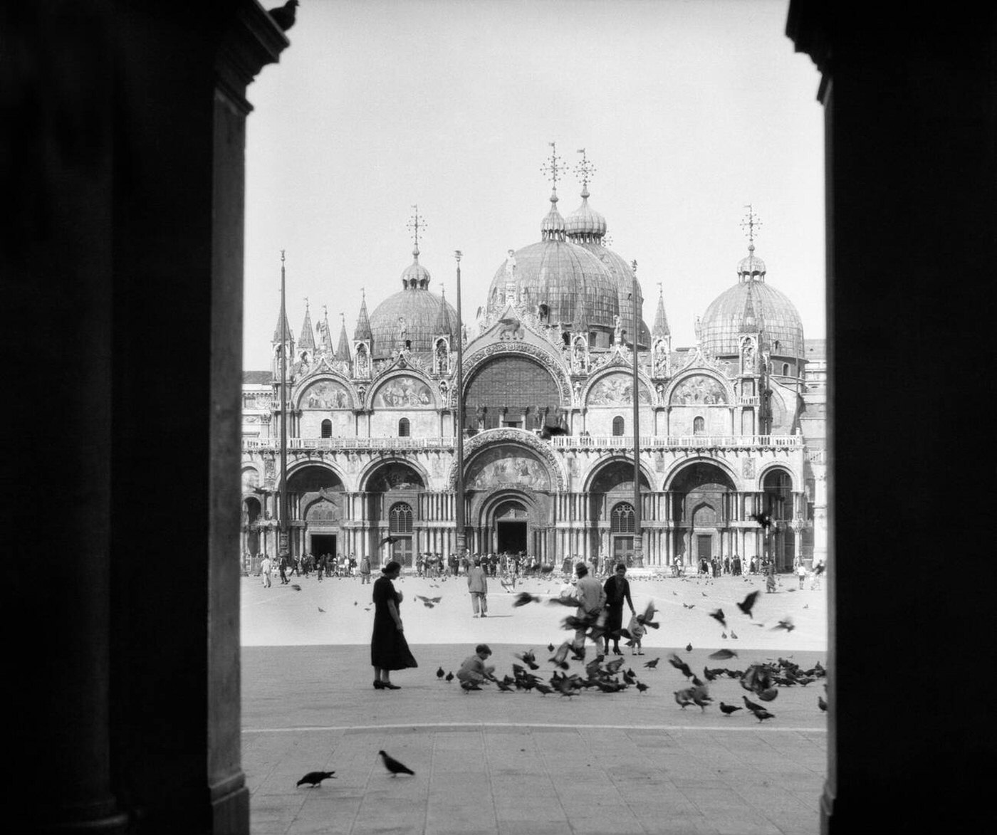 View through doorway to people feeding pigeons in front of St. Mark's Cathedral, Venice, Italy, 1920s.