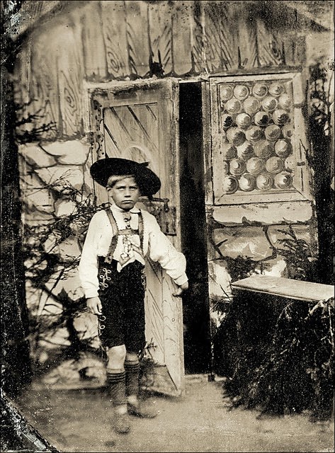 A Bavarian boy appears disappointed when trying to peek behind a mysterious door backdrop, Germany, early 20th century.