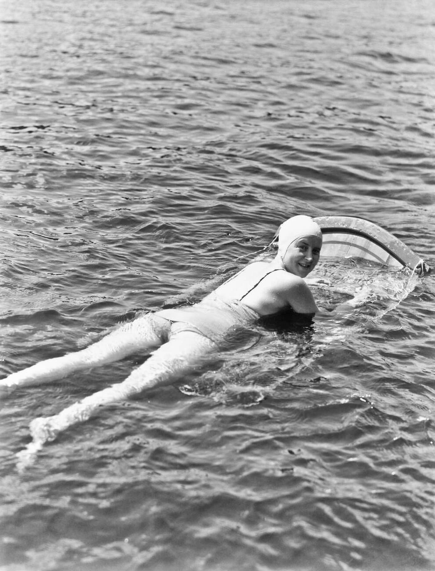 Woman wearing a bathing cap and one-piece suit lying on an aqua ski board looking at the camera over her shoulder at Muskoka Lake, Canada, 1935.