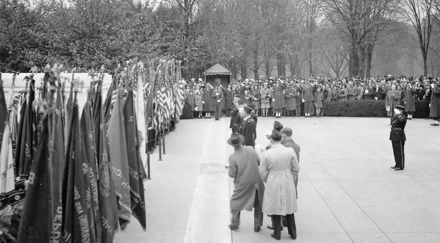 Photographers, uniformed military personnel, and civilian spectators observe a military contingent saluting a long row of flags, likely in Ohio during celebrations marking the end of World War II, 1945.