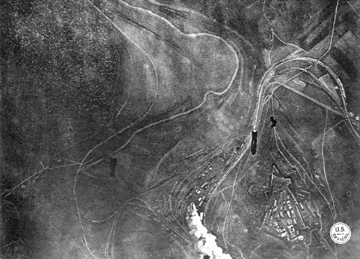 Bombing Montmedy, 42 km north of Verdun, while American troops advance in the Meuse-Argonne sector; three bombs have been released by a U.S. bomber, one striking a supply station, the other two in mid-air, with black puffs of smoke indicating anti-aircraft fire.