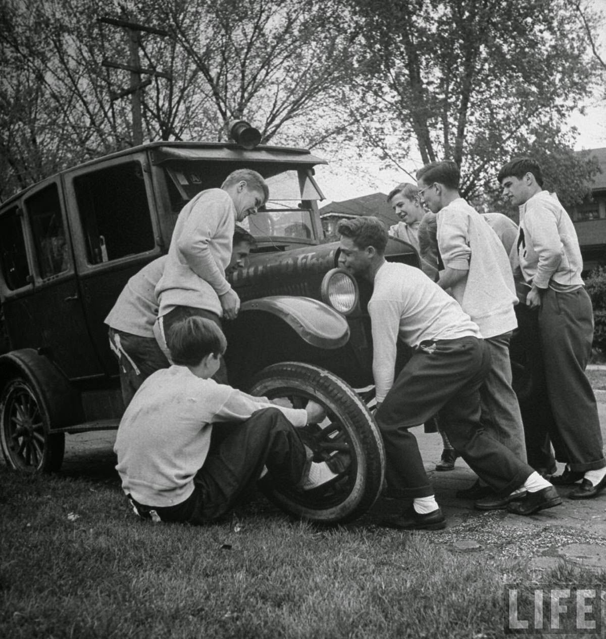 Teenaged boys, who are junior members of the Molesters Club (as indicated by large "M"s sewn on the seats of their pants) rather than using a jack, simply lift the car so that the one pulling on tire can change it.