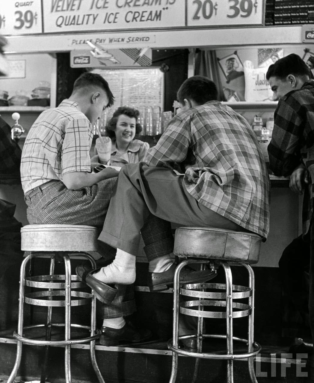 Young men in plaid shirts drinking ice-cream sodas at soda fountain.