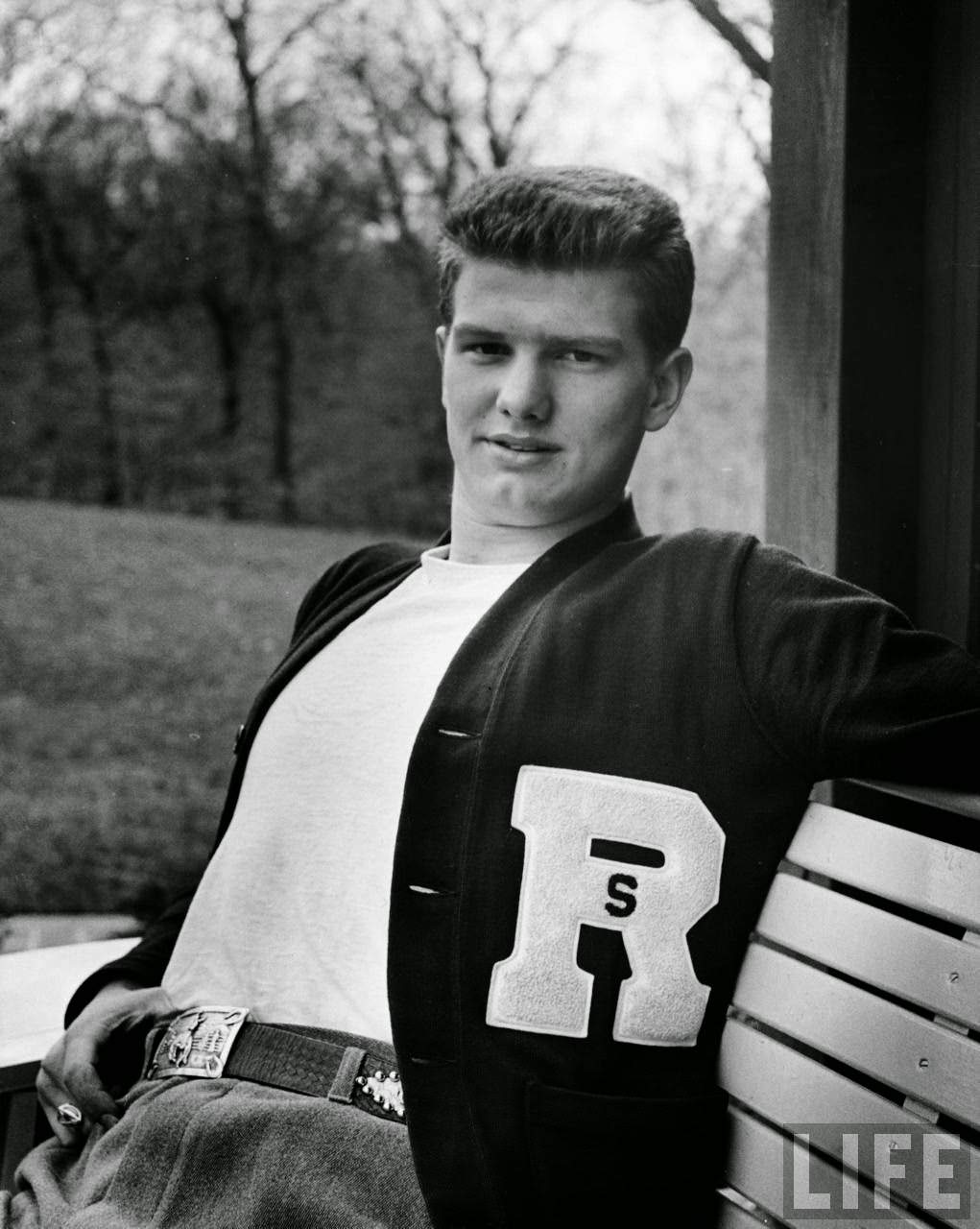Teenage boy wearing sweater w. letter R, relaxing outside.