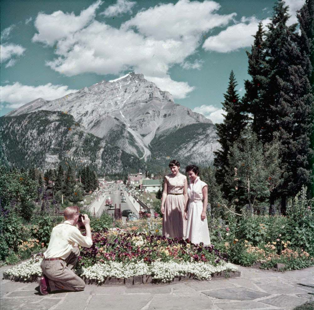 A man photographs two women in the garden of the Administration Building, with the main street of the town of Banff behind, 1952.