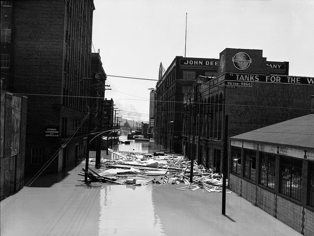 View of Hardware Company, John Deere, and other buildings on a flooded street; a large pile of debris floats in the water, 1951