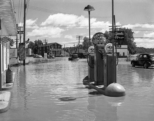 Flooding on a busy street with a gas station in the foreground, Beck's Grocery in the background, and several cars attempting to drive through water, 1951