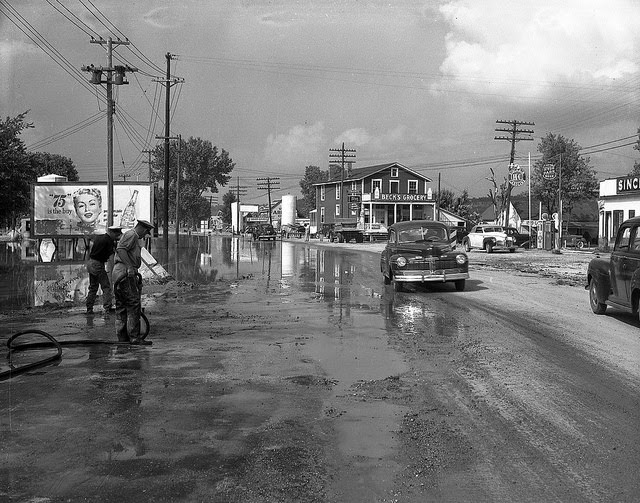 Flood damage on Rt. 63-54 shows receded water on a busy street in Cedar City, with Beck's Grocery store in the background, 1951