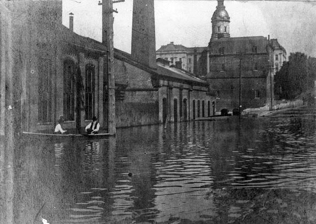 Two men float in a boat along a flooded street in Jefferson City, Missouri, with the old State Capitol building in the background, 1903