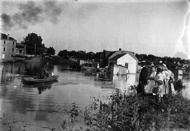 A group stands on dry land as a boat navigates a flooded area in Jefferson City, Missouri, 1906
