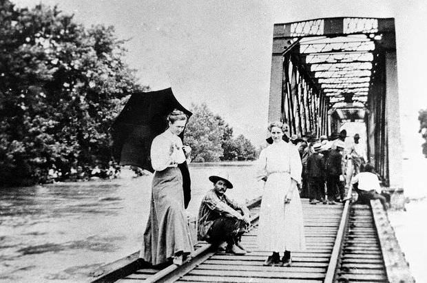 Two women and a man stand on a flooded railroad bridge, with a large group of people gathered behind them, 1909