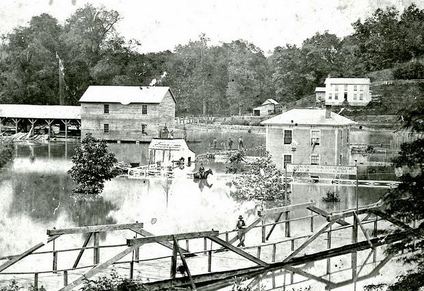 View of flooding in Tuscumbia, Missouri, with a bridge in the foreground and buildings in the back; several men, including one on horseback, survey the damage, 1926
