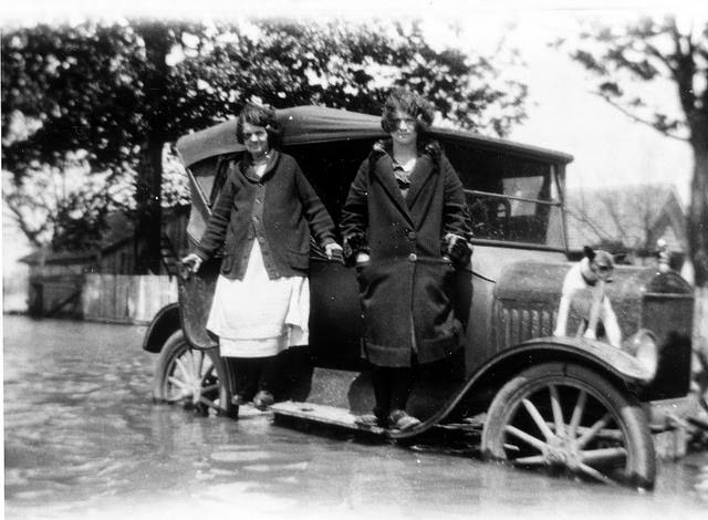 Two women and a dog stand atop an automobile mostly submerged in flood waters, 1937