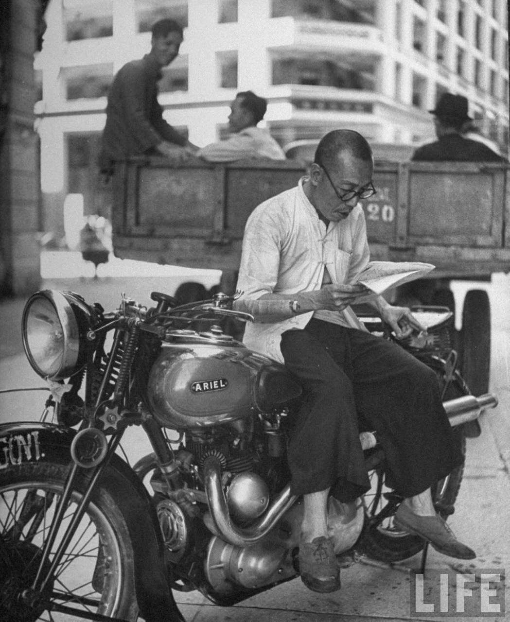 Chinese man sitting on a motorcycle, reading a newspaper.